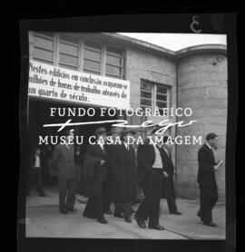 Grupo de pessoas junto a placa com lema empresarial. Cooperativa dos Pedreiros, rua da Alegria.